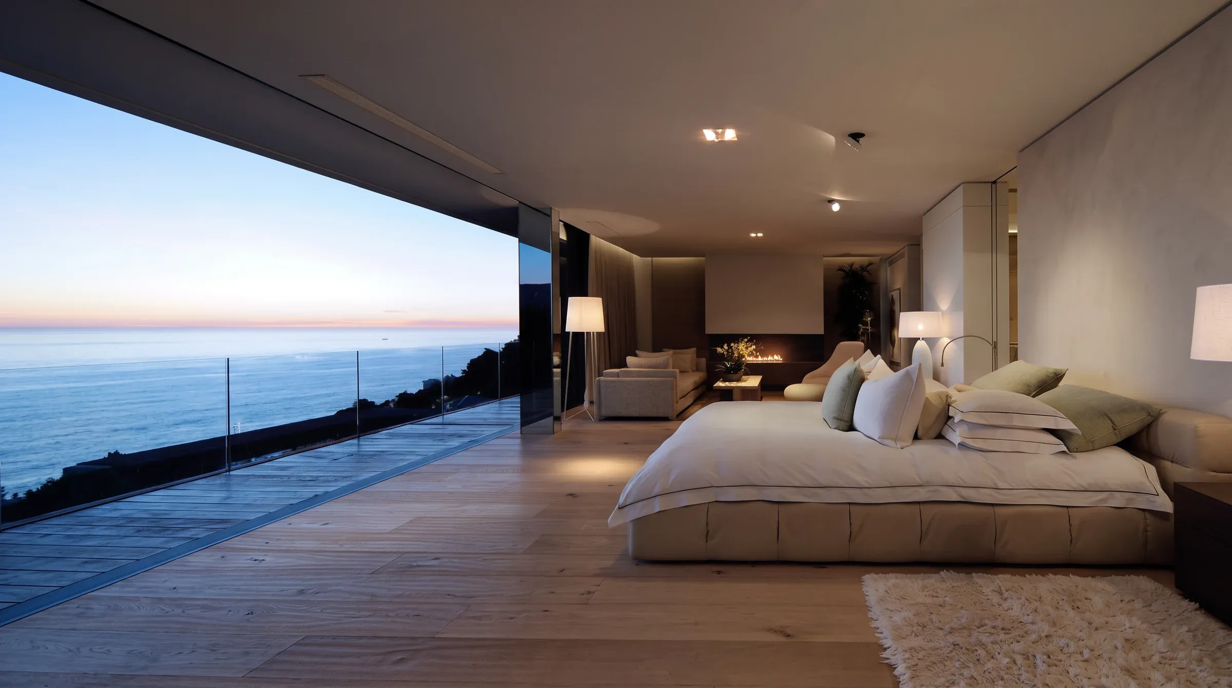 A contemporary villa bedroom at dusk, the bed set sideways, a vast picture window open to the sea.