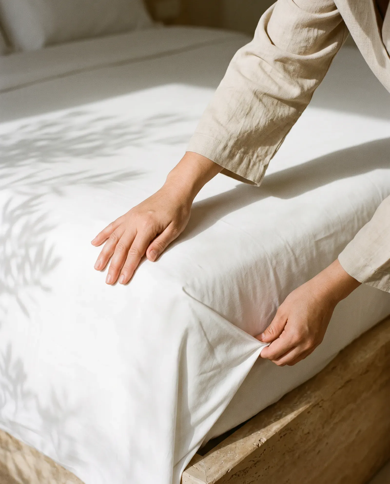 Hands smoothing a white sheet over a bed in the morning light.